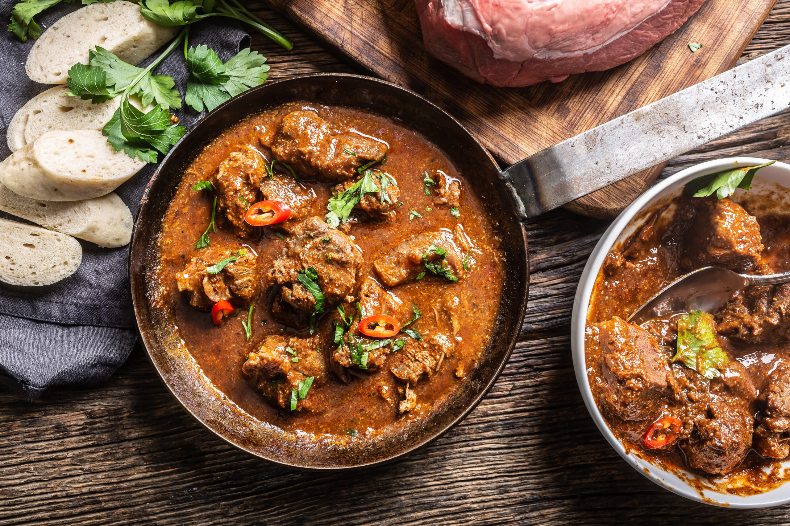 Beef or pork goulash in a pan and in a bowl next to dumplings viewed from the top.